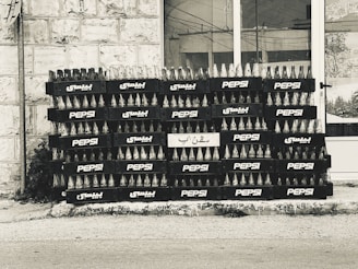 A stack of empty glass bottles organized neatly in black crates bearing the Pepsi logo and Arabic script. The crates are arranged in several rows and columns against a tiled building facade.