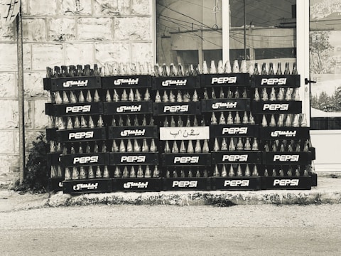 A stack of empty glass bottles organized neatly in black crates bearing the Pepsi logo and Arabic script. The crates are arranged in several rows and columns against a tiled building facade.