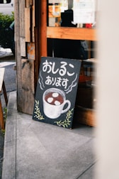 A chalkboard sign leaning against a wooden building. The sign features Japanese text and a drawing of a steaming cup filled with a brown liquid and white circular objects, likely representing a beverage. The bottom of the board is decorated with simple green leafy designs. The background includes part of the building and a glimpse of the street.