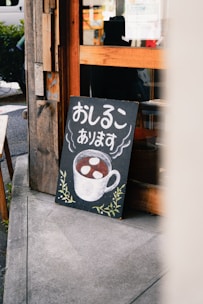 A chalkboard sign leaning against a wooden building. The sign features Japanese text and a drawing of a steaming cup filled with a brown liquid and white circular objects, likely representing a beverage. The bottom of the board is decorated with simple green leafy designs. The background includes part of the building and a glimpse of the street.
