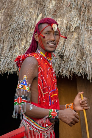 A person wearing traditional Masai attire adorned with intricate beadwork and accessories. The background features a thatched roof, indicating a rural or cultural setting. The attire is predominantly red with colorful patterns.