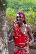 A person dressed in traditional attire with red fabric adorned with intricate beadwork and decorations. They are holding a stick and standing outdoors near a large tree, with lush green foliage in the background.