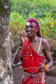 A person dressed in traditional attire with red fabric adorned with intricate beadwork and decorations. They are holding a stick and standing outdoors near a large tree, with lush green foliage in the background.