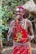A person is wearing traditional attire decorated with colorful beads and patterns, consisting of a red cloth adorned with white and silver embellishments. They have accessories including a headpiece with a circular ornament hanging down the forehead. The background features lush green foliage and a thatched structure.