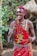 A person is wearing traditional attire decorated with colorful beads and patterns, consisting of a red cloth adorned with white and silver embellishments. They have accessories including a headpiece with a circular ornament hanging down the forehead. The background features lush green foliage and a thatched structure.