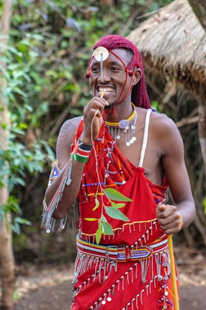 A person is wearing traditional attire decorated with colorful beads and patterns, consisting of a red cloth adorned with white and silver embellishments. They have accessories including a headpiece with a circular ornament hanging down the forehead. The background features lush green foliage and a thatched structure.