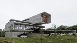 A modern architectural structure with a minimalist design featuring large glass windows and a cantilevered upper section. The building is set amidst a lush green landscape with trees scattered around. The exterior is predominantly made of concrete with sleek finishes. There is a white car parked on a driveway beside the building.