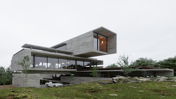 A modern architectural structure with a minimalist design featuring large glass windows and a cantilevered upper section. The building is set amidst a lush green landscape with trees scattered around. The exterior is predominantly made of concrete with sleek finishes. There is a white car parked on a driveway beside the building.