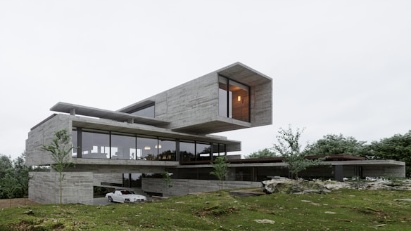 A modern architectural structure with a minimalist design featuring large glass windows and a cantilevered upper section. The building is set amidst a lush green landscape with trees scattered around. The exterior is predominantly made of concrete with sleek finishes. There is a white car parked on a driveway beside the building.