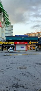 A street view featuring several businesses next to each other, including a cheese store, a Vodafone store, and a pasta restaurant. A white car is parked near the stores. There are signs in bright colors on the storefronts. The sky is partly cloudy, with a prominent palm frond visible at the top left.