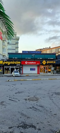 A street view featuring several businesses next to each other, including a cheese store, a Vodafone store, and a pasta restaurant. A white car is parked near the stores. There are signs in bright colors on the storefronts. The sky is partly cloudy, with a prominent palm frond visible at the top left.