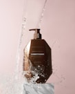 A brown bottle of Moroccan Argan Oil conditioner is positioned on a white pedestal against a soft pink background. Water splashes around the bottle, giving a dynamic and refreshing feel.