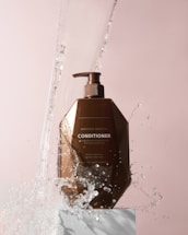 A brown bottle of Moroccan Argan Oil conditioner is positioned on a white pedestal against a soft pink background. Water splashes around the bottle, giving a dynamic and refreshing feel.