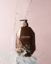 A brown bottle of Moroccan Argan Oil conditioner is positioned on a white pedestal against a soft pink background. Water splashes around the bottle, giving a dynamic and refreshing feel.
