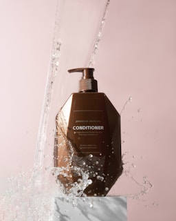 A brown bottle of Moroccan Argan Oil conditioner is positioned on a white pedestal against a soft pink background. Water splashes around the bottle, giving a dynamic and refreshing feel.