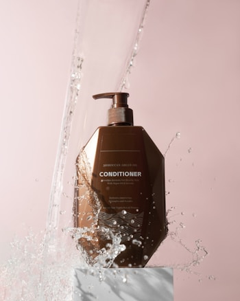 A brown bottle of Moroccan Argan Oil conditioner is positioned on a white pedestal against a soft pink background. Water splashes around the bottle, giving a dynamic and refreshing feel.