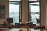 An office space with a large wooden desk positioned in front of two wide windows. Outside the windows, a scenic view of a large body of water, a distant landscape featuring buildings, and a bridge can be seen. On the desk, there are several items including papers, a phone, and office supplies. The room is minimally decorated, with a few pieces of wall art.