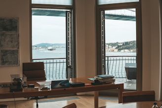 An office space with a large wooden desk positioned in front of two wide windows. Outside the windows, a scenic view of a large body of water, a distant landscape featuring buildings, and a bridge can be seen. On the desk, there are several items including papers, a phone, and office supplies. The room is minimally decorated, with a few pieces of wall art.