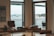 An office space with a large wooden desk positioned in front of two wide windows. Outside the windows, a scenic view of a large body of water, a distant landscape featuring buildings, and a bridge can be seen. On the desk, there are several items including papers, a phone, and office supplies. The room is minimally decorated, with a few pieces of wall art.