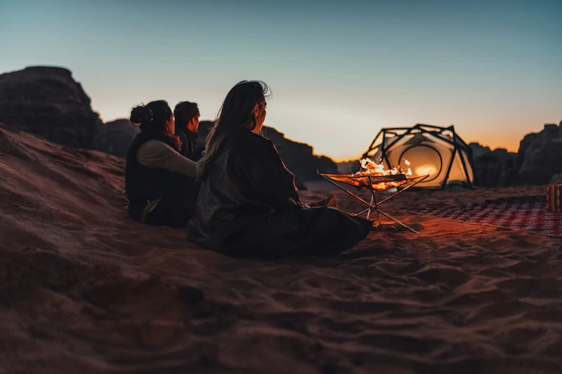 A fire burning in a raised pit on the sand in front of a lit camp