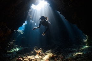 A person is swimming underwater in a sunlit section of a cave. Sunlight beams down through the water, illuminating the rocky seabed and surrounding coral formations.