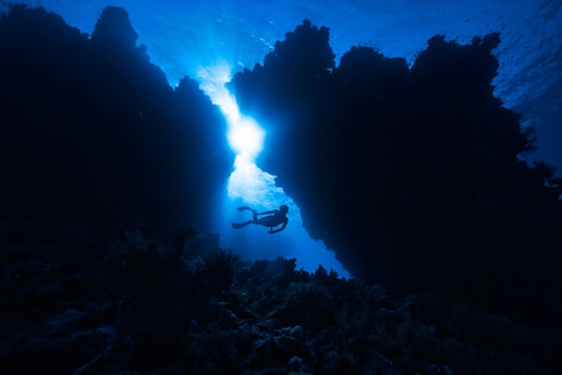 A diver is exploring an underwater coral landscape, with vivid sunlight streaming through a gap in the rocky formations, creating a dramatic silhouette against the deep blue water.