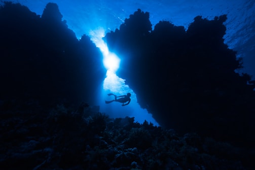 A diver is exploring an underwater coral landscape, with vivid sunlight streaming through a gap in the rocky formations, creating a dramatic silhouette against the deep blue water.