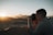 A person is taking a photograph of a desert landscape during sunset, with the sunlight casting warm tones over the sand dunes. The scene captures the silhouette of distant mountains and a clear sky.