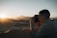 A person is taking a photograph of a desert landscape during sunset, with the sunlight casting warm tones over the sand dunes. The scene captures the silhouette of distant mountains and a clear sky.