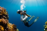 A diver is underwater, wearing a snorkeling mask and fins, holding a camera. They are surrounded by vibrant coral formations, and sunlight filters through the clear blue water overhead creating a serene atmosphere.