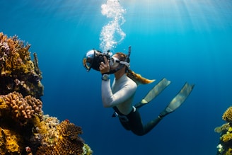 A diver is underwater, wearing a snorkeling mask and fins, holding a camera. They are surrounded by vibrant coral formations, and sunlight filters through the clear blue water overhead creating a serene atmosphere.