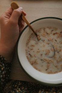 A hand holding a spoon is seen poised over a bowl containing a milky liquid with grains or cereal floating on the surface. The bowl is placed on a wooden table, and the person's arm is partially visible, wearing a patterned, possibly floral, garment.