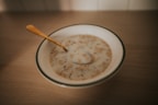 A bowl of cereal or porridge with milk sitting on a wooden table. The cereal appears to be mixed with grains and nuts. A golden spoon rests inside the bowl.