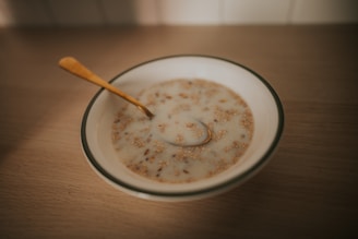 A bowl of cereal or porridge with milk sitting on a wooden table. The cereal appears to be mixed with grains and nuts. A golden spoon rests inside the bowl.