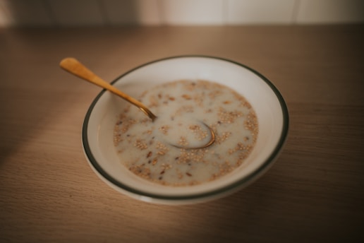 A bowl of cereal or porridge with milk sitting on a wooden table. The cereal appears to be mixed with grains and nuts. A golden spoon rests inside the bowl.