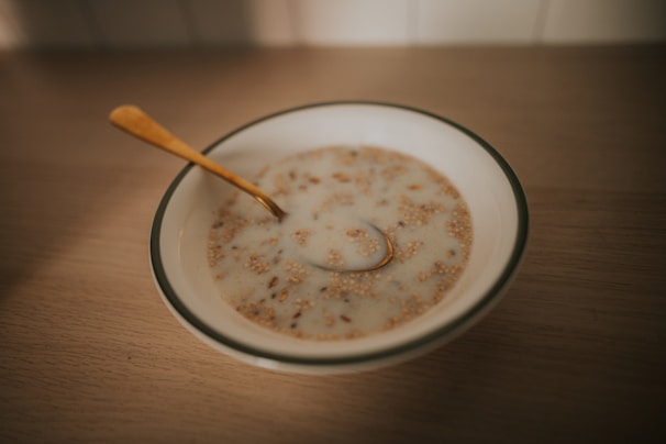 A bowl of cereal or porridge with milk sitting on a wooden table. The cereal appears to be mixed with grains and nuts. A golden spoon rests inside the bowl.
