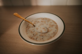 A bowl of cereal or porridge with milk sitting on a wooden table. The cereal appears to be mixed with grains and nuts. A golden spoon rests inside the bowl.