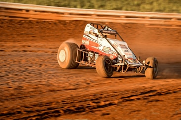 A race car is speeding on a dirt track, creating a cloud of dust behind it. The vehicle is designed for racing on an oval track, featuring robust tires and a protective frame over the cockpit. The background is blurred, indicating high speed.