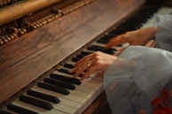 Hands are playing on a piano, with a focus on the wooden texture of the piano and the soft lighting giving a warm ambiance. The hands are delicately positioned over the keys, suggesting a gentle and thoughtful interaction with the instrument.