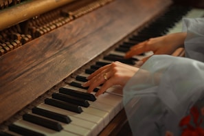 Hands are playing on a piano, with a focus on the wooden texture of the piano and the soft lighting giving a warm ambiance. The hands are delicately positioned over the keys, suggesting a gentle and thoughtful interaction with the instrument.