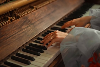 Hands are playing on a piano, with a focus on the wooden texture of the piano and the soft lighting giving a warm ambiance. The hands are delicately positioned over the keys, suggesting a gentle and thoughtful interaction with the instrument.