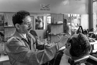 A barber is carefully shaving a client's face in a well-lit barbershop. The shop contains shelves with various items, and mirrors are mounted on the wall. Both the barber and the client appear focused, with additional figures reflected in the mirrors.