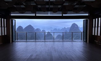 A spacious, empty room with large sliding glass doors open to a balcony, offering a panoramic view of mist-shrouded mountains. The interior features dark wooden flooring and slatted window coverings, and the overall atmosphere is serene and tranquil.