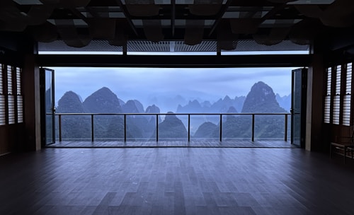 A spacious, empty room with large sliding glass doors open to a balcony, offering a panoramic view of mist-shrouded mountains. The interior features dark wooden flooring and slatted window coverings, and the overall atmosphere is serene and tranquil.
