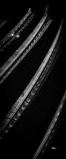 An aerial view of multiple highways or roads lit by streetlights and depicted in high contrast with dark shadows accentuating the curves of the roads. Sparse traffic is visible with a few cars and possibly pedestrians, giving a sense of movement and infrastructure.