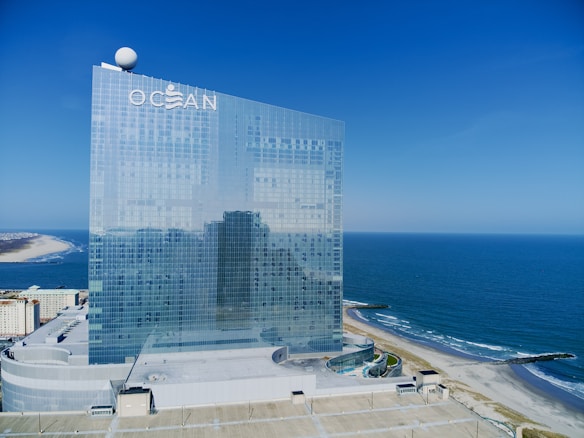 A tall, modern glass building stands next to a beach with the ocean stretching into the distance. The building has the word 'OCEAN' at the top, with a spherical object on one corner. Below, a sandy beach curves along the shoreline.