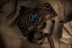 Two intricately carved dark wood candle holders feature elephant designs with a single blue flower-shaped candle placed within one. They are surrounded by crumpled brown paper and burlap fabric, set on a wooden surface, creating a rustic and earthy composition.