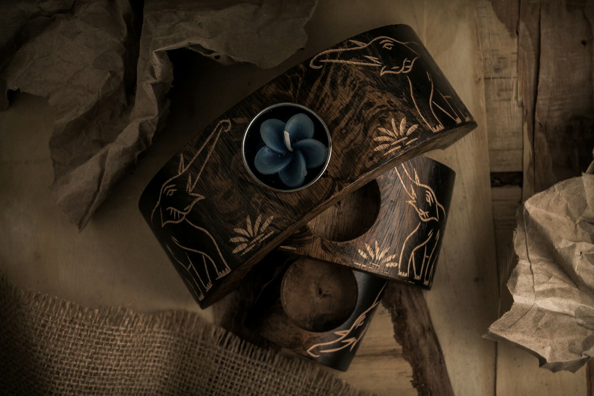 Two intricately carved dark wood candle holders feature elephant designs with a single blue flower-shaped candle placed within one. They are surrounded by crumpled brown paper and burlap fabric, set on a wooden surface, creating a rustic and earthy composition.
