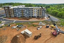 A construction site with a multi-story residential building in the background, surrounded by greenery. Heavy machinery, including a crane and a bulldozer, are present along with various construction materials and equipment scattered across the dirt-covered ground. A container is also visible on the site.