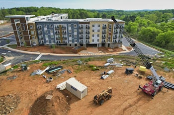 A construction site with a multi-story residential building in the background, surrounded by greenery. Heavy machinery, including a crane and a bulldozer, are present along with various construction materials and equipment scattered across the dirt-covered ground. A container is also visible on the site.